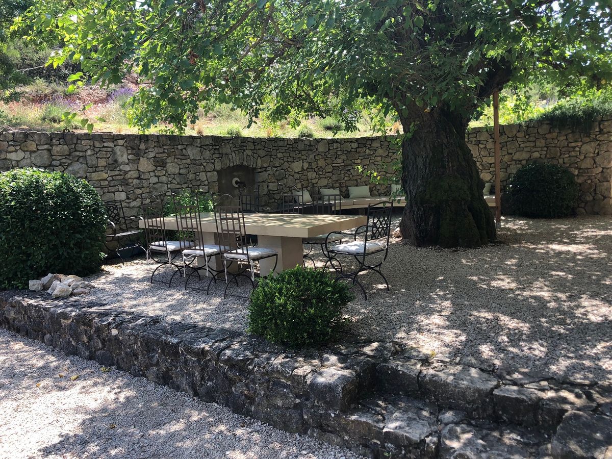 La cour à l’est : la table de pierre, les chaises en fer forgé et le tronc du mûrier, contre le mur de pierre sèche.
