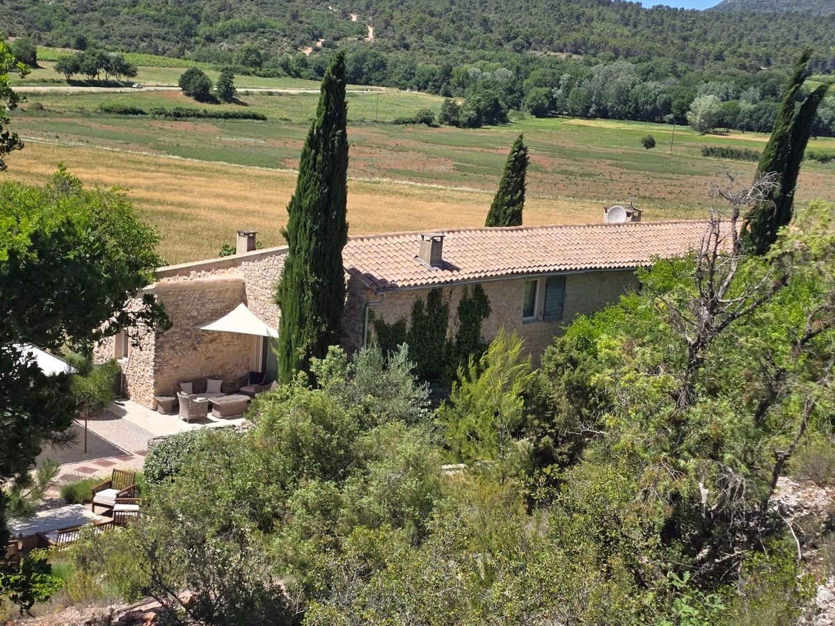 Le mas vu de la colline, entre cyprès et chênes, avec les champs et les collines derrière.