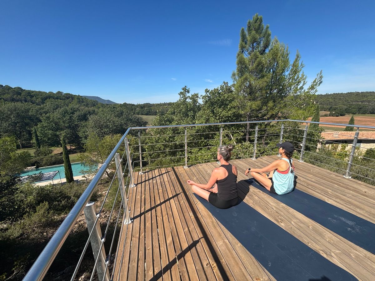 La plateforme en bois au-dessus de la piscine : deux personnes en séance de yoga face aux collines boisées.