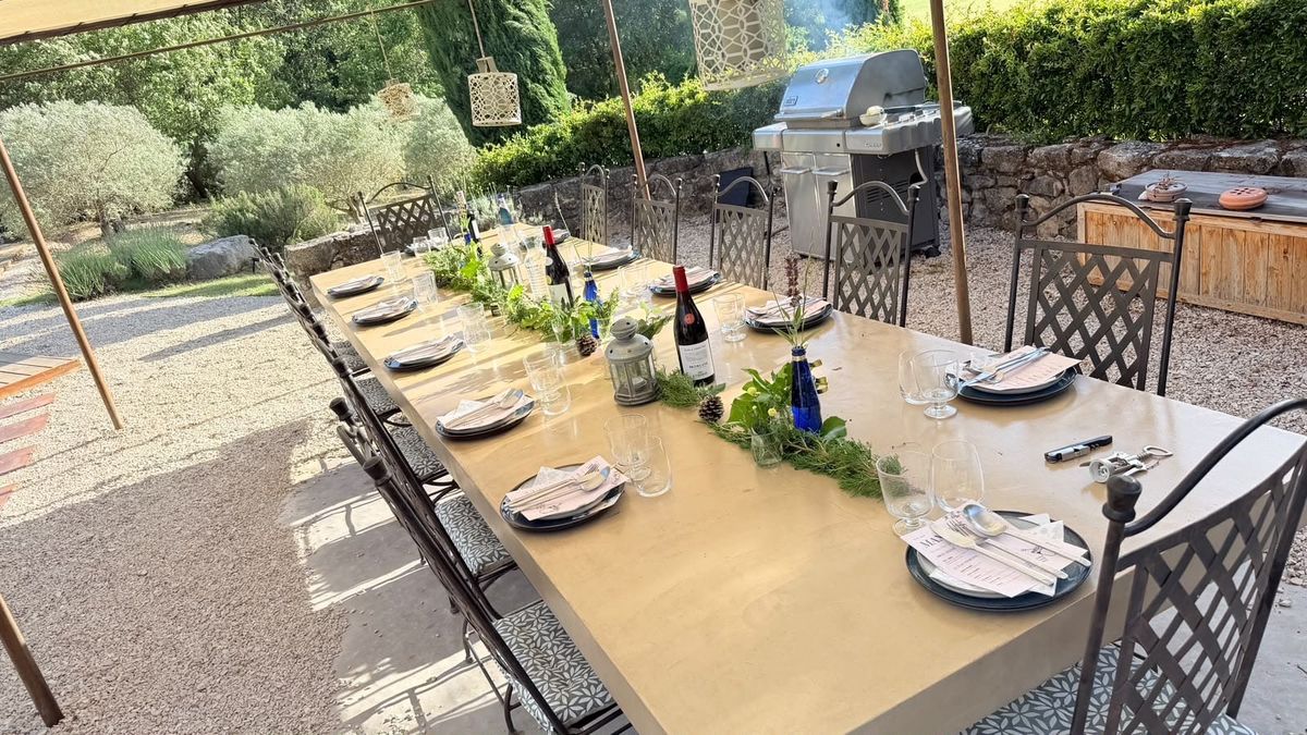 Une longue table dressée en plein air, avec bouteilles de vin, couverts et bouquets.